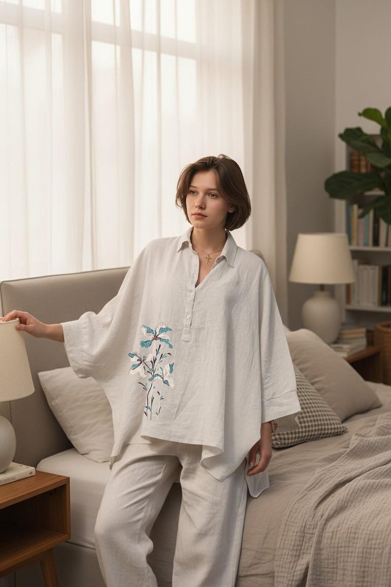 May include: A light-colored linen pajama set. The top is a loose-fitting, button-down shirt with a floral embroidery design in shades of blue and white. The pants are a matching, relaxed fit. The set is displayed in a bedroom setting.