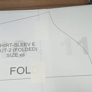 May include: A white paper shirt sleeve pattern with the text "SHIRT-SLEEVE E CUT-2 (FOLDED) SIZE xs" and the word "FOLD". The pattern is taped to a wooden surface.