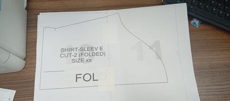 May include: A white paper shirt sleeve pattern with the text "SHIRT-SLEEVE E CUT-2 (FOLDED) SIZE xs" and the word "FOLD". The pattern is taped to a wooden surface.