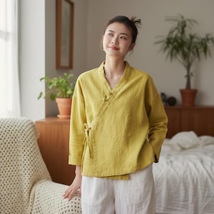May include: A mustard yellow wrap top with a V-neck and long sleeves, paired with white linen pants. The top features a side tie closure and decorative buttons. The outfit is casual and comfortable.