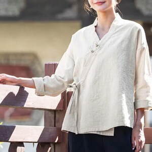May include: Off-white linen wrap top with a V-neck and long sleeves. The top features a tie closure at the waist and is paired with black pants. The style is reminiscent of traditional Asian clothing.