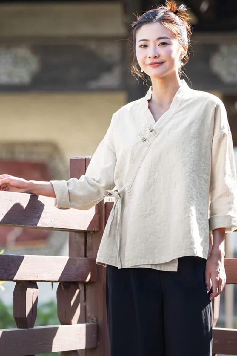 May include: Off-white linen wrap top with a V-neck and long sleeves. The top features a tie closure at the waist and is paired with black pants. The style is reminiscent of traditional Asian clothing.
