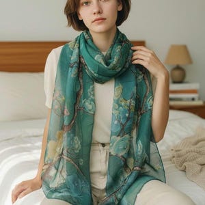 May include: A teal scarf with a floral design featuring branches and flowers. The scarf is draped around the neck and shoulders. The scarf is made of a lightweight material.