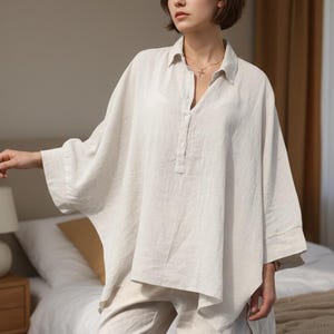 May include: A light beige linen shirt and matching pants. The shirt has a relaxed fit with a button-down collar and three buttons at the neckline. The sleeves are three-quarter length. The pants are a straight-leg style.