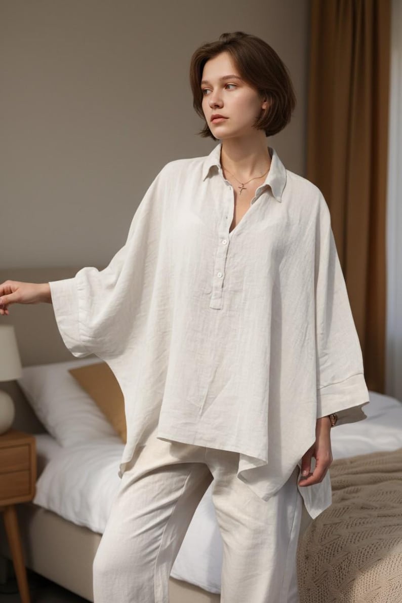 May include: A light beige linen shirt and matching pants. The shirt has a relaxed fit with a button-down collar and three buttons at the neckline. The sleeves are three-quarter length. The pants are a straight-leg style.