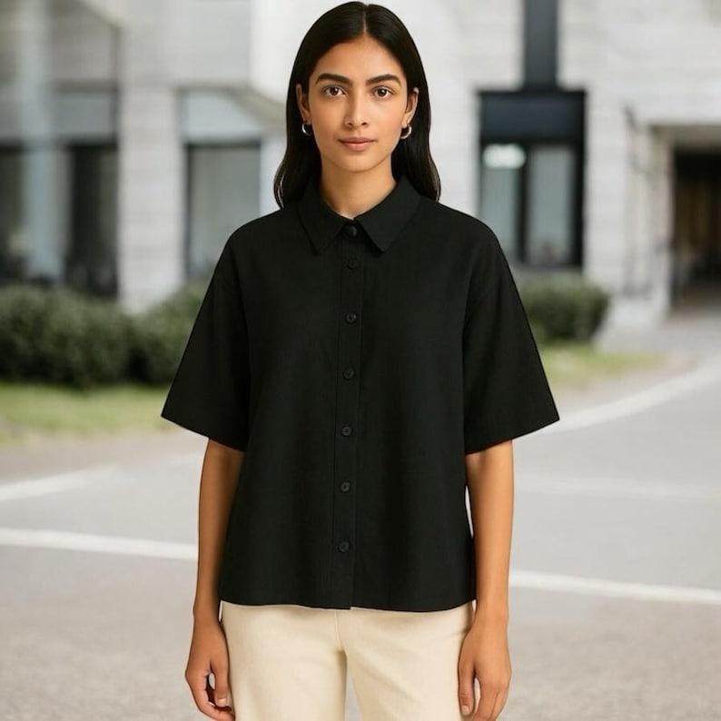 May include: A black short-sleeved shirt with a button-down front and a collared neckline. The shirt is worn with light-coloured trousers. The background shows a building.