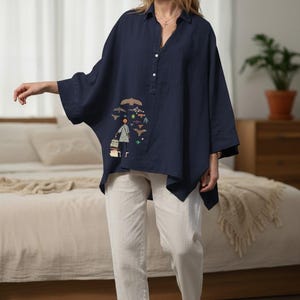 May include: A navy blue, loose-fitting blouse with a unique embroidered design featuring a person and birds. The blouse has a button-down front and three-quarter sleeves. Paired with white pants, creating a casual, artistic look.