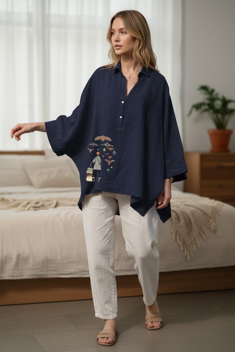 May include: A navy blue, loose-fitting blouse with a unique embroidered design featuring a person and birds. The blouse has a button-down front and three-quarter sleeves. Paired with white pants, creating a casual, artistic look.
