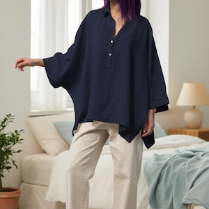 May include: A navy blue, oversized linen shirt with a V-neck and button details is paired with cream-colored jeans. The outfit is completed with white canvas sneakers. The model is standing in a bedroom setting.