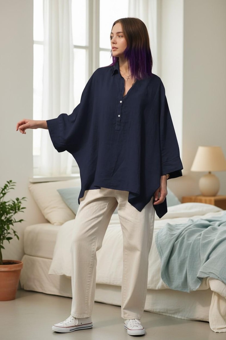 May include: A navy blue, oversized linen shirt with a V-neck and button details is paired with cream-colored jeans. The outfit is completed with white canvas sneakers. The model is standing in a bedroom setting.