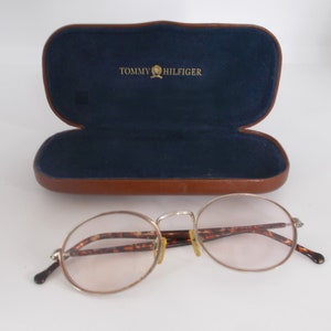 May include: Silver-rimmed eyeglasses with oval lenses and tortoise-shell arms rest in front of an open brown leather case with a blue interior. The case is embossed with the words "TOMMY HILFIGER" in gold.
