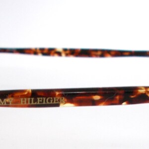 May include: Close-up of Tommy Hilfiger eyeglasses. The glasses have a round frame and brown and tan tortoise shell arms. The brand name is printed in gold on the arm.