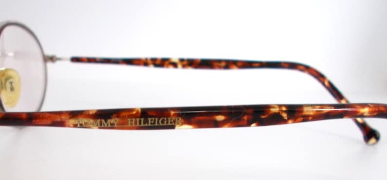 May include: Close-up of Tommy Hilfiger eyeglasses. The glasses have a round frame and brown and tan tortoise shell arms. The brand name is printed in gold on the arm.