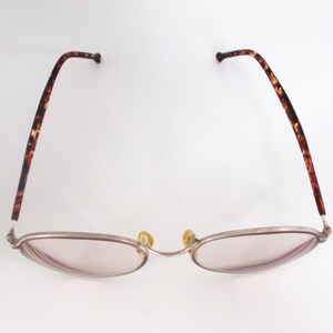 May include: Oval-shaped eyeglasses with a silver frame and pink-tinted lenses. The temples feature a brown tortoiseshell pattern. The nose pads are yellow. The glasses are designed for vision correction.