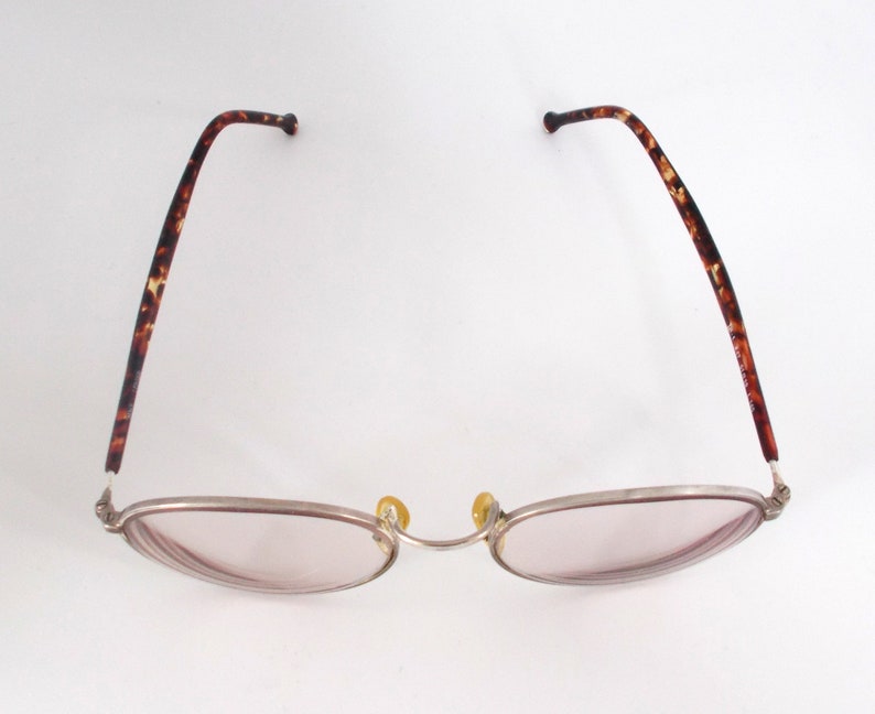 May include: Oval-shaped eyeglasses with a silver frame and pink-tinted lenses. The temples feature a brown tortoiseshell pattern. The nose pads are yellow. The glasses are designed for vision correction.