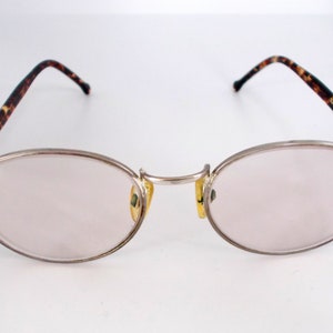 May include: Oval-shaped eyeglasses with a silver metal frame and light pink lenses. The temples feature a brown and black tortoiseshell pattern. The nose pads are yellow. The glasses are designed for vision correction.