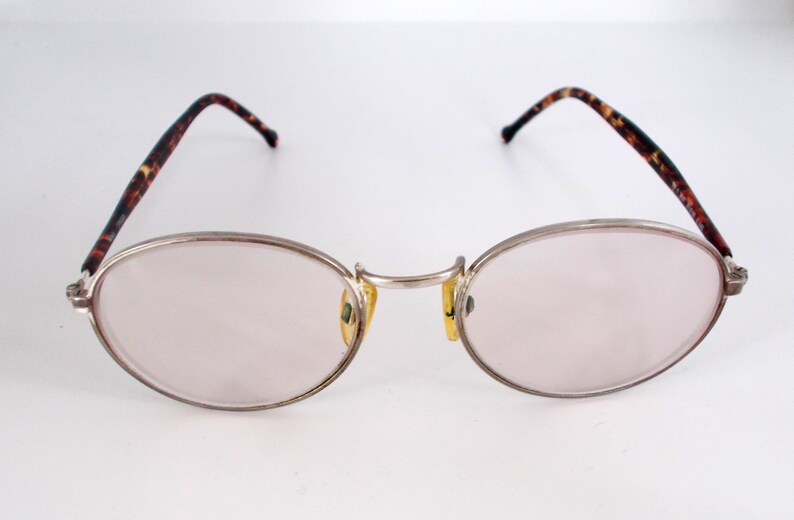 May include: Oval-shaped eyeglasses with a silver metal frame and light pink lenses. The temples feature a brown and black tortoiseshell pattern. The nose pads are yellow. The glasses are designed for vision correction.