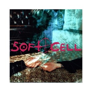 May include: Album cover art for Soft Cell. The image features the band's name in pink, overlaid on a dark, textured background with a crosshair symbol. Two hands are visible in the foreground, with a patterned surface.
