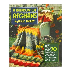 May include: A book cover titled "A Rainbow of Afghans" by Leslie Linsley. The cover features a colorful chevron-patterned afghan draped over a white wicker chair. The afghan has red, orange, yellow, green, and blue sections. Text on the cover states "With More than 70 Full-Color Variations to Crochet and Knit."