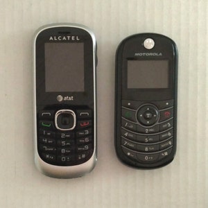 May include: Two cell phones. The phone on the left is an Alcatel with the AT&T logo. The phone on the right is a Motorola. Both phones are black with silver accents and have keypads with numbers and letters.