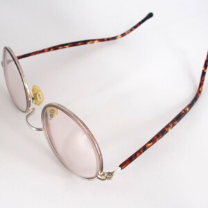 May include: Oval-shaped eyeglasses with a silver frame and pink-tinted lenses. The temples feature a brown and black tortoiseshell pattern. The nose pads are yellow. The glasses are set against a white background.