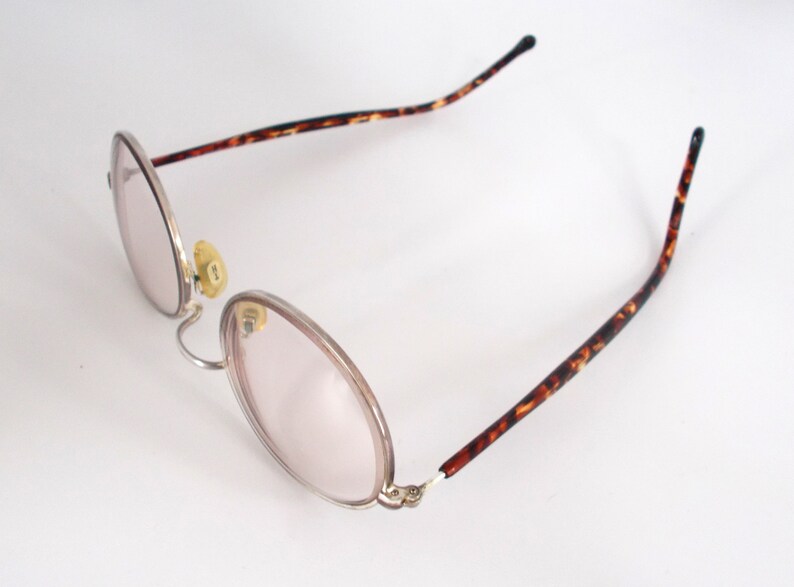 May include: Oval-shaped eyeglasses with a silver frame and pink-tinted lenses. The temples feature a brown and black tortoiseshell pattern. The nose pads are yellow. The glasses are set against a white background.