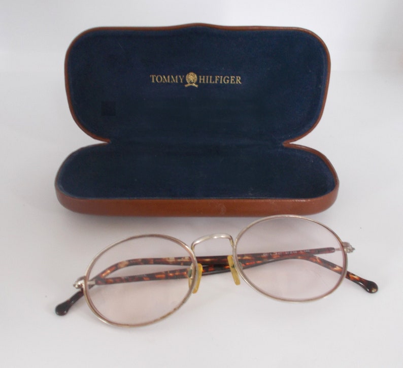 May include: Silver-rimmed eyeglasses with oval lenses and tortoise-shell arms rest in front of an open brown leather case with a blue interior. The case is embossed with the words "TOMMY HILFIGER" in gold.