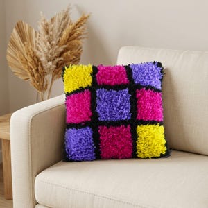 May include: A square throw pillow featuring a shaggy, tufted texture with a grid pattern of nine squares. The squares are colored in bright yellow, hot pink, and purple, separated by thick black yarn lines throughout the entire design.