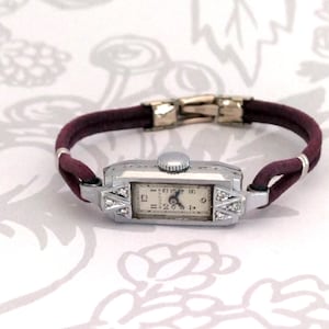 May include: Vintage Bulova wristwatch with a rectangular silver-tone case and a cream-colored face. The watch features diamond accents and a burgundy leather band. The clasp is gold-toned. A classic timepiece.
