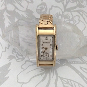 May include: A vintage Bulova wristwatch with a rectangular gold-tone case and a matching expandable band. The watch face is off-white with black numerals and hands. The watch measures approximately 0.75 inches wide.