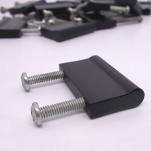 May include: Black rectangular furniture hardware with two silver screws. The hardware has a curved top and is likely used for attaching or securing furniture components. Several similar pieces are out of focus in the background.