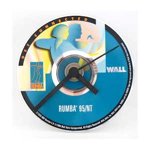 May include: A round clock with a blue face featuring the text "RUMBA 95/NT" and a graphic of two silhouetted figures. The clock hands are black, and the clock is mounted on a white background. The clock also has the text "GET CONNECTED" and "WALL DATA".