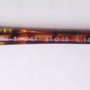 May include: Close-up of a brown and yellow tortoiseshell eyeglass arm. The arm has the following text: TM-1 #47-51 19 L.145. The arm is a dark brown color with yellow and brown marbling.