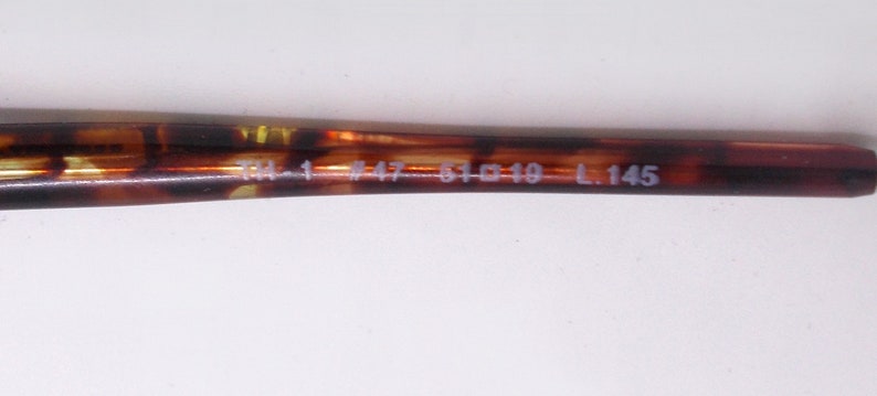 May include: Close-up of a brown and yellow tortoiseshell eyeglass arm. The arm has the following text: TM-1 #47-51 19 L.145. The arm is a dark brown color with yellow and brown marbling.