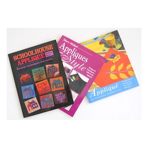 May include: Three books on a white surface. The book on the left is titled "Schoolhouse Applique" with colorful house illustrations. The middle book is "Appliques Style" by Mary Mulari. The book on the right is "Appliqué" by Lucinda Ganderton.