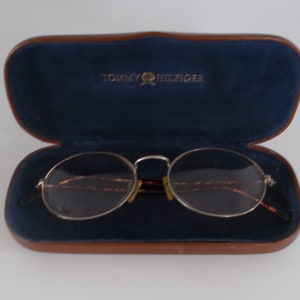 May include: Open brown leather eyeglasses case with a navy blue interior and the Tommy Hilfiger logo in gold. Silver-rimmed eyeglasses with oval lenses rest inside the case.