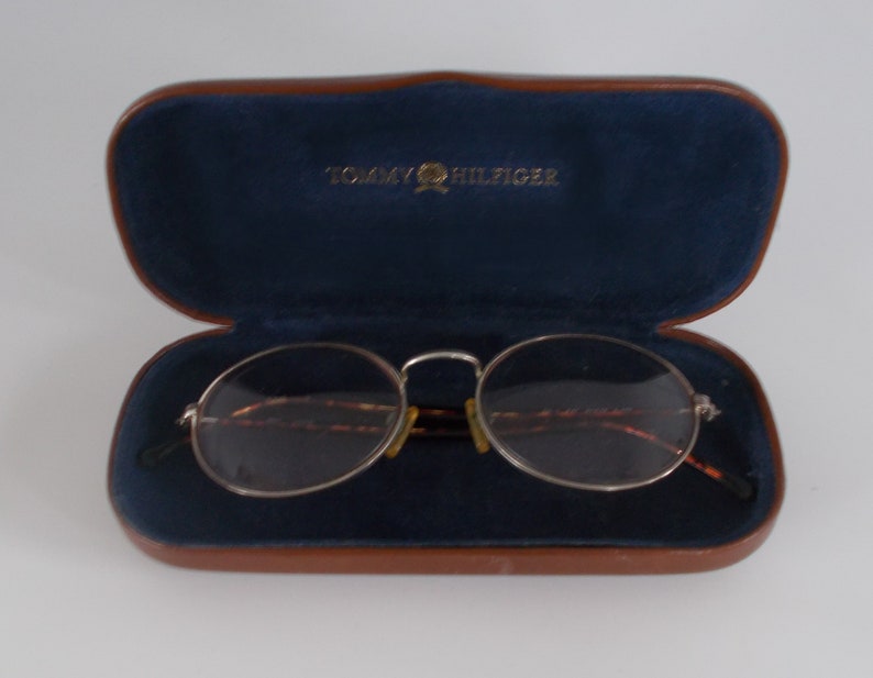 May include: Open brown leather eyeglasses case with a navy blue interior and the Tommy Hilfiger logo in gold. Silver-rimmed eyeglasses with oval lenses rest inside the case.