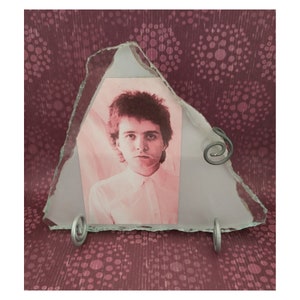 May include: A unique, triangular glass photo frame with a jagged edge. The frame holds a sepia-toned portrait of a person in a white shirt. The frame is supported by two silver metal stands and a decorative swirl.