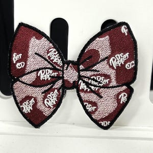 May include: A burgundy and pink bow-tie patch with the Dr. Pepper logo embroidered in white. The bow-tie has a black border and is set against a white background.