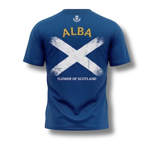 May include: Blue t-shirt with a white distressed Scottish flag design on the back. The word "ALBA" is printed in gold above the flag, with "FLOWER OF SCOTLAND" below. A thistle emblem is at the top.