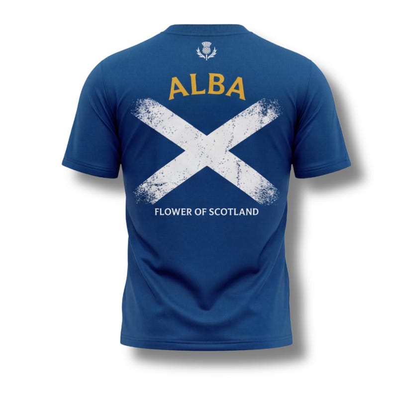 May include: Blue t-shirt with a white distressed Scottish flag design on the back. The word "ALBA" is printed in gold above the flag, with "FLOWER OF SCOTLAND" below. A thistle emblem is at the top.