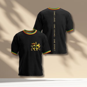 Op de afbeelding: Zwarte t-shirts met een geel, rood en groen ontwerp. Eén shirt heeft een leeuwenembleem en de andere de tekst "ONE LOVE • ONE SOUND • ONE SPIRIT" verticaal gedrukt. De kraag en mouwen hebben een rood, geel en groene rand.