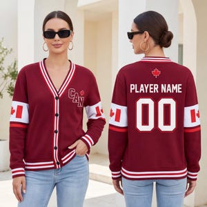 May include: A maroon V-neck cardigan with white and maroon striped trim. The front features the text CAN and a maple leaf. The back displays the text PLAYER NAME, the number 00, a maple leaf, and white sleeve bands with red maple leaf icons.