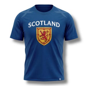May include: A blue t-shirt with the word "SCOTLAND" in white block letters above a shield. The shield features a red lion rampant on a gold background. The shirt has short sleeves and a crew neck.
