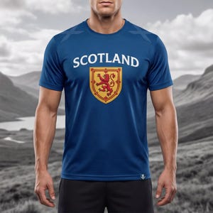 May include: A blue t-shirt with the word "SCOTLAND" in white block letters above a shield emblem. The emblem features a red lion rampant on a gold background. The shirt is short-sleeved.