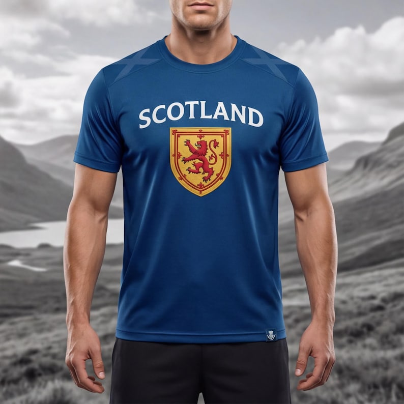 May include: A blue t-shirt with the word "SCOTLAND" in white block letters above a shield emblem. The emblem features a red lion rampant on a gold background. The shirt is short-sleeved.