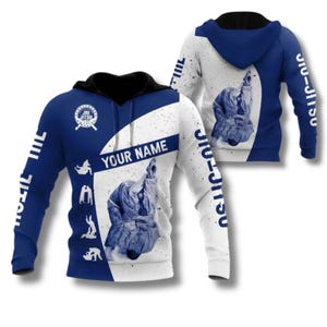 Può includere: Una felpa con cappuccio blu e bianca con cappuccio e coulisse neri. Il design include una grafica di due persone che praticano jiu-jitsu, la scritta "JIU JITSU" sulle maniche, un logo circolare e uno spazio per "YOUR NAME".