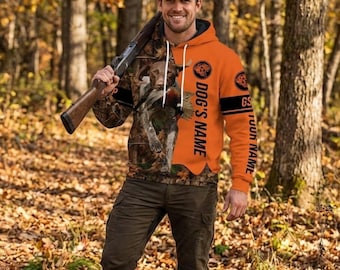 May include: An orange and camouflage hoodie with the text "DOG'S NAME" and "YOUR NAME" printed on the sleeves. The hoodie features a hunting dog and pheasant design. The person in the image is wearing a camouflage baseball cap and holding a rifle.
