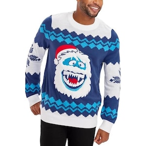 Op de afbeelding: Een blauw-witte kersttrui met een cartoon yeti-gezicht met een rode kerstmuts. De trui heeft een blauw zigzagpatroon en witte sneeuwvlokontwerpen op de mouwen en schouders.