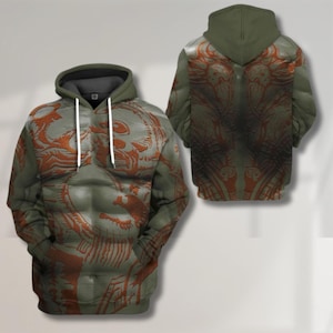 May include: Olive green hoodie with a detailed orange design on the front and back. The front features a stylized figure, while the back showcases a symmetrical pattern. The hoodie has a drawstring hood and a kangaroo pocket.